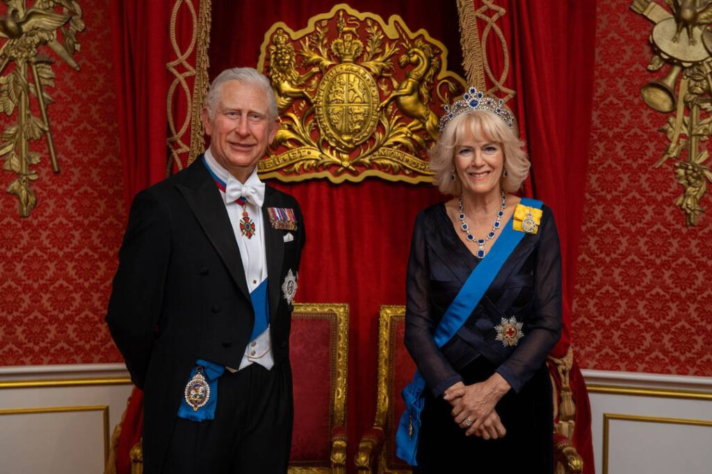 Wax figures of King Charles III and Queen Camilla at Madame Tussauds London, posed in a royal setting with red and gold decor