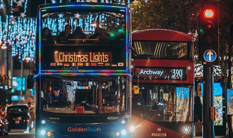 Golden Tours open-top bus lit up for a London Christmas Lights tour, driving past festive decorations alongside a red London bus.