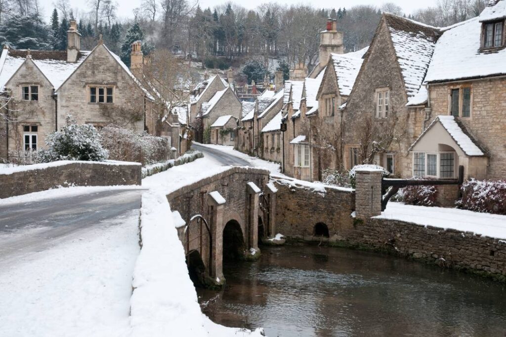 Traditional stone cottages in a Cotswolds village covered in snow with a stone bridge over a river, a classic English countryside winter scene.