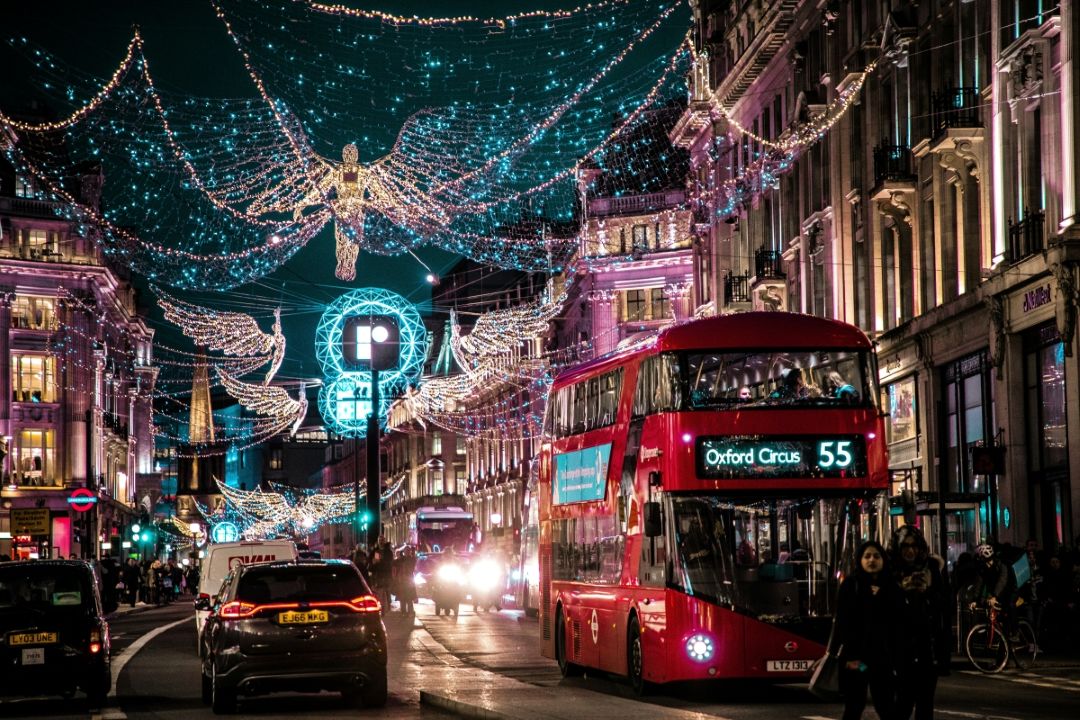 Oxford Street in London decorated with glowing Christmas lights and angel displays, with a red bus and black cabs passing underneath.